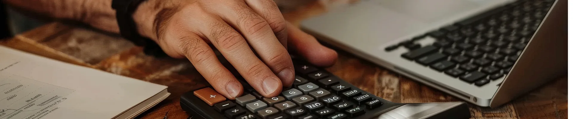الشخص الذي يستخدم آلة حاسبة في مكتب خشبي مع كمبيوتر محمول وهاتف ذكي ومستندات مالية.